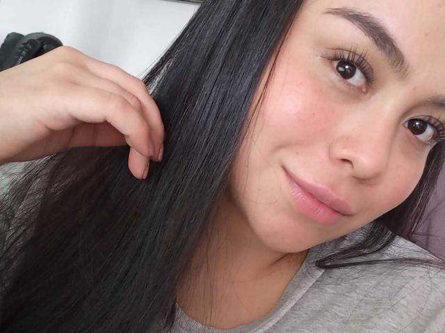 MartinaMarX with long black hair, holding her hair with her right hand, has a slight smile, brown eyes, and pinkish lips, wearing a grey shirt, looking cute.
