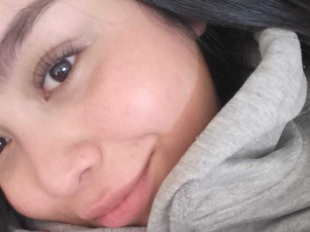 MartinaMarX's face with light brown skin, dark eyes, and a small smile, wearing a grey hoodie, showing a slight tan line on her cheek. Her black hair is visible.