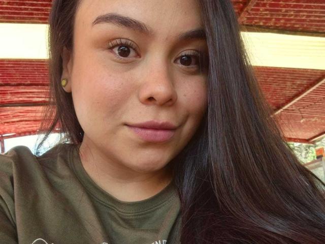 MartinaMarX has medium brown skin, long straight dark brown hair, small yellow earbuds, and is wearing a green shirt with her pouty lips slightly closed, brown eyes looking at camera.