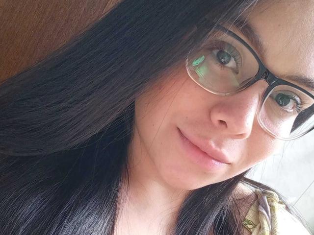 O rosto de MartinaMarX, com cabelos pretos longos, usando óculos e com um leve sorriso. Ela tem pele marrom clara e um nariz pequeno, com seu olhar verde visível..