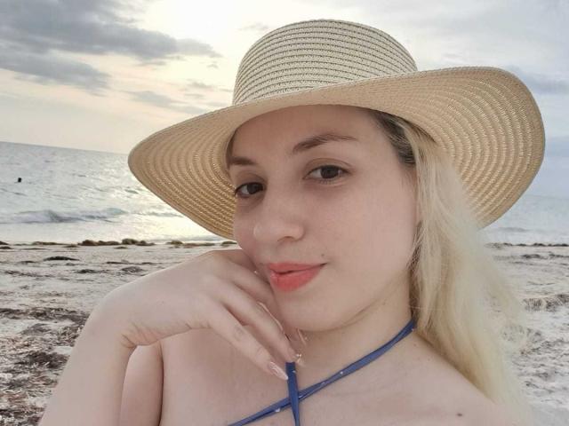 HaileyDavies with pale skin, blonde hair, wearing a straw hat and blue halter top, has her hand near her pink lips, and is looking at the camera.