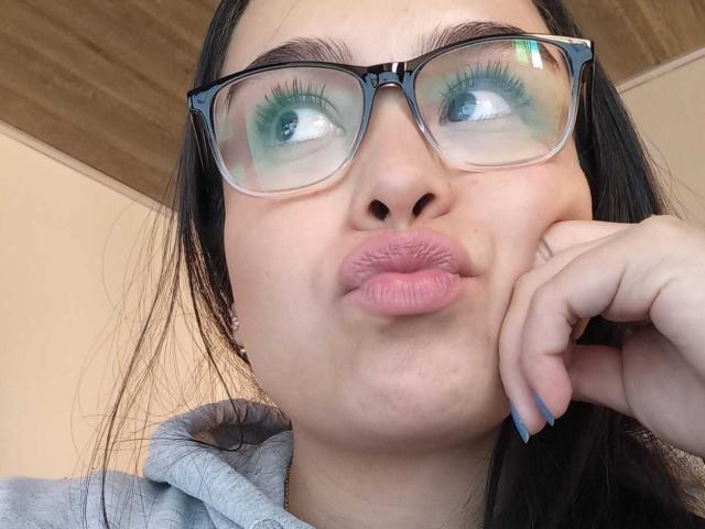 MartinaMarX with pouty pink lips, black-framed glasses, green eyeliner, and blue nail polish, biting her finger, looking up, wearing a gray hoodie.