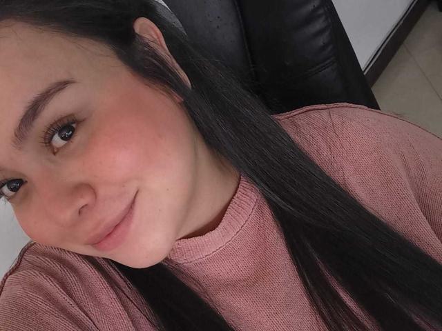 MartinaMarX tem cabelos pretos longos, pele rosada e olhos castanhos. Ela está vestindo um suéter rosa, com um leve sorriso no rosto, e seus seios não estão visíveis..
