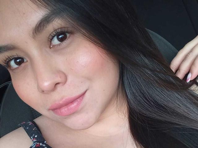 MartinaMarX has a cute face with brown eyes, pink lips, and straight black hair. She's smiling softly, showing a bit of her cheek and wearing a floral dress.