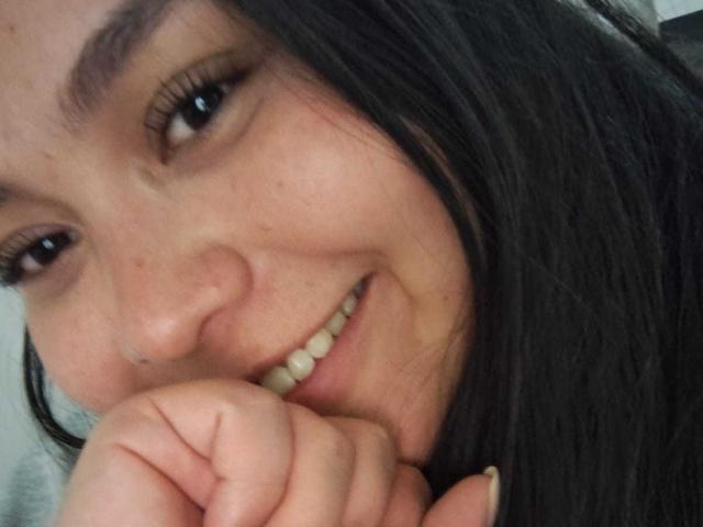 MartinaMarX with dark hair, smiling, showing teeth, and resting her chin on her hand, her nose is slightly upturned and she has a small nose piercing.