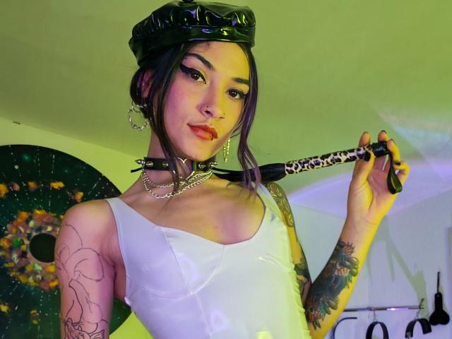 AkiraNishimura69 with dark hair in a shiny black cap, white tank top, choker, holding a black-and-white chain whip, tattooed arms, and orange lipstick, looking sexy.