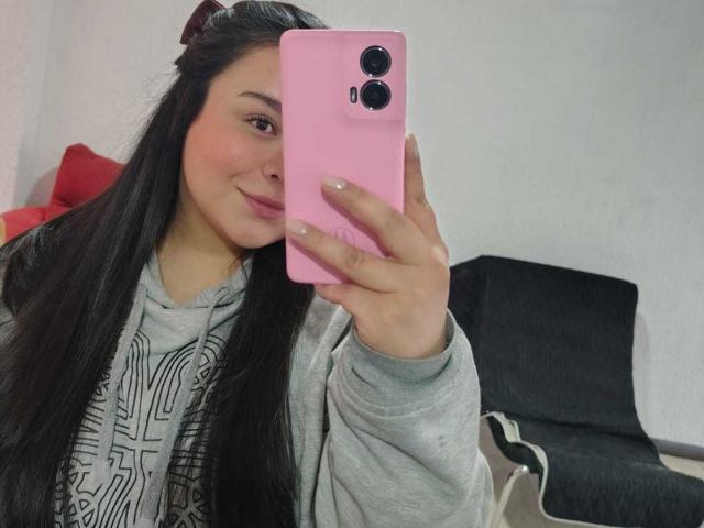 MartinaMarX with long black hair, pink phone, grey hoodie, smiling at mirror, holding phone with black camera, pink case, in a room with red and black furniture.