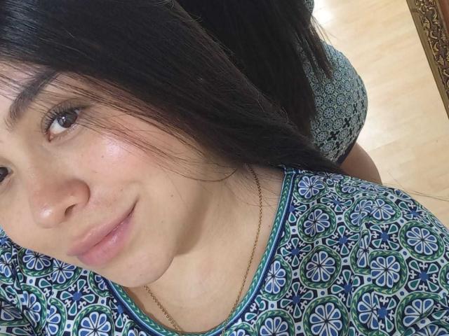 MartinaMarX hat ein hübsches Gesicht mit braunen Augen, schwarzen Haaren und einem leichten Lächeln. Sie trägt ein blaues, mit Blumen gemustertes Hemd, das ihre Brüste zeigt, und eine goldene Halskette..