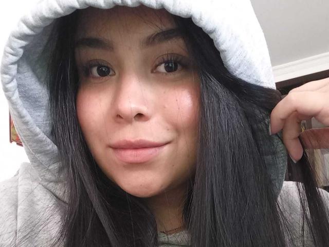 O rosto de MartinaMarX, com cabelos pretos longos, olhos castanhos e um leve sorriso, vestindo um moletom cinza com o capuz levantado, segurando um fio de seu cabelo..