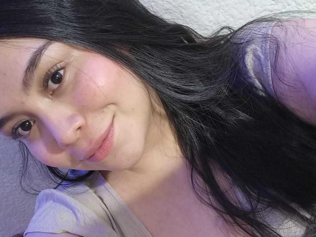 MartinaMarX has long black hair, a slight smile, pink cheeks, and brown eyes. She's wearing a grey shirt, lying down, with soft purple lighting on her face.