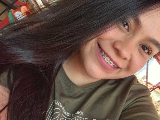 O rosto sorridente de MartinaMarX, com cabelo preto liso, olhos castanhos e aparelho metálico nos dentes. Ela está vestindo uma camisa verde, que mostra seu traseiro bonito e atraente..