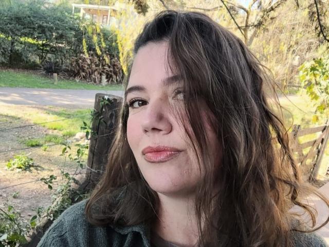 SophieHotSweet with messy brown hair, pinkish lips, and a slight smirk, wearing a green jacket, in a sunlit outdoor setting, with a wooden fence in the background.