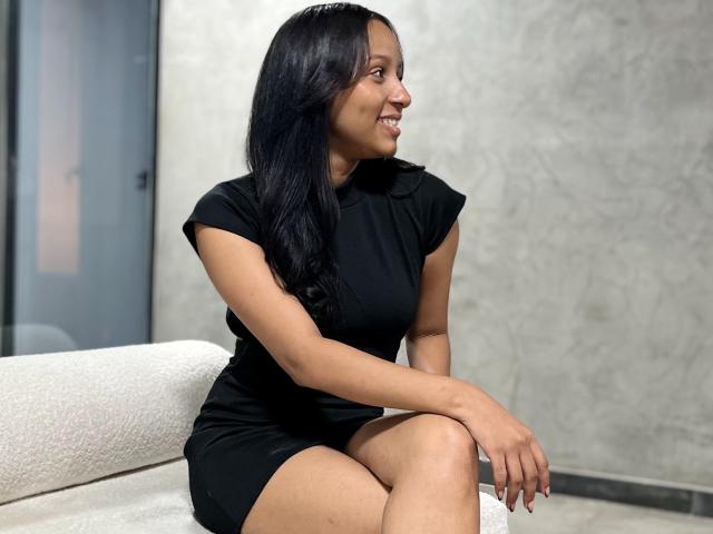 RaquelZafd, sitzend auf einem weißen Sofa, trägt ein schwarzes Kleid, hat langes, schwarzes Haar, lächelt, hat die Beine gekreuzt, wobei ihr Hintern zu sehen ist, und ihre Hände ruhen auf ihren Knien..
