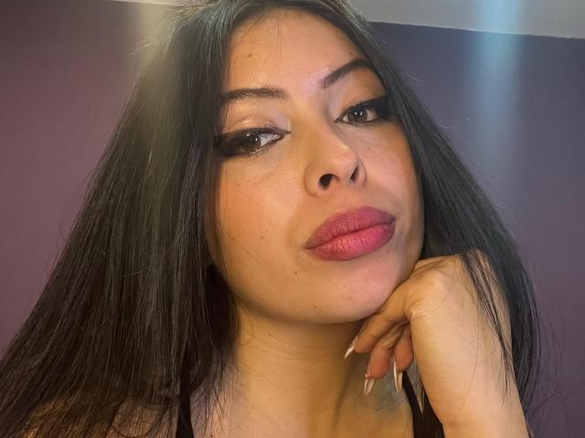 KateWelsin with long black hair, full pink lips, and dark eyeliner, resting her chin on her hand, looking at the camera with a slightly pouty expression.