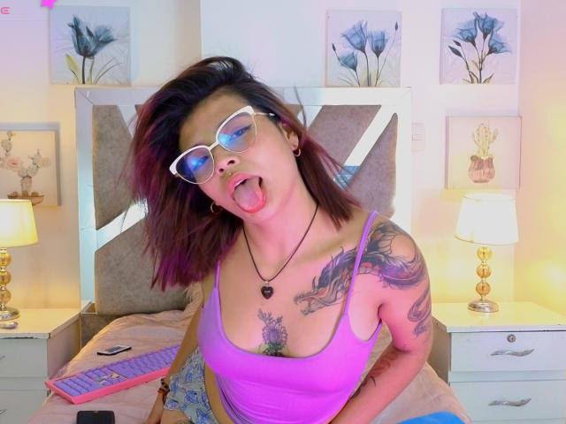 LanaFlowers69 with glasses, tongue out, purple tank top, tattoos on shoulders and chest, holding pink keyboard, brown hair, white bed, gold lamps, flower paintings, and white nightstands.