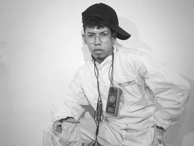 LianStonne with glasses, wearing a backwards cap, white jacket, and ID lanyard, sitting with a serious expression, hands on knees, in a black-and-white photo.