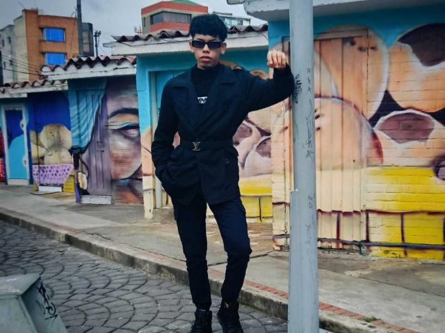 LianStonne is a slim dude with black hair, wearing dark sunglasses, a black shirt, black pants, and black boots, standing by a graffiti-covered wall, leaning on a pole.