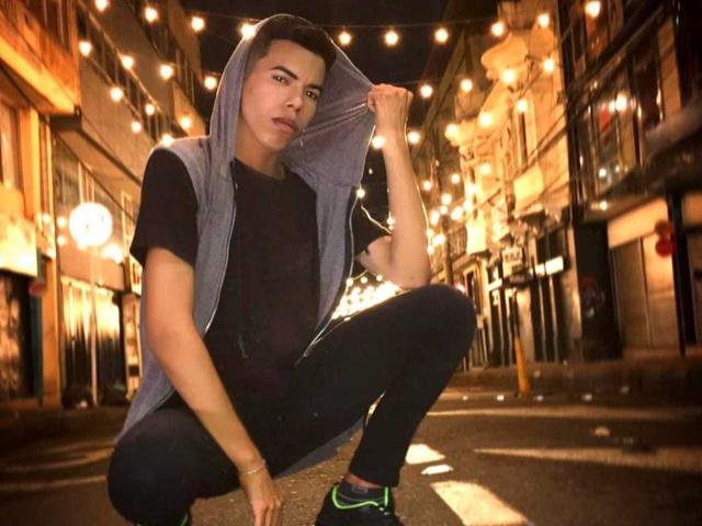 LianStonne, squatting, holding grey hoodie, black shirt, black pants, green sneakers, lights overhead, night, short dark hair, slight pout, left hand on knee, right hand on hoodie.