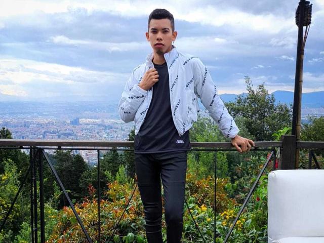 LianStonne stands in a white jacket with black lettering, black shirt, and black pants, leaning on a metal railing, with a cloudy sky and greenery in the background.
