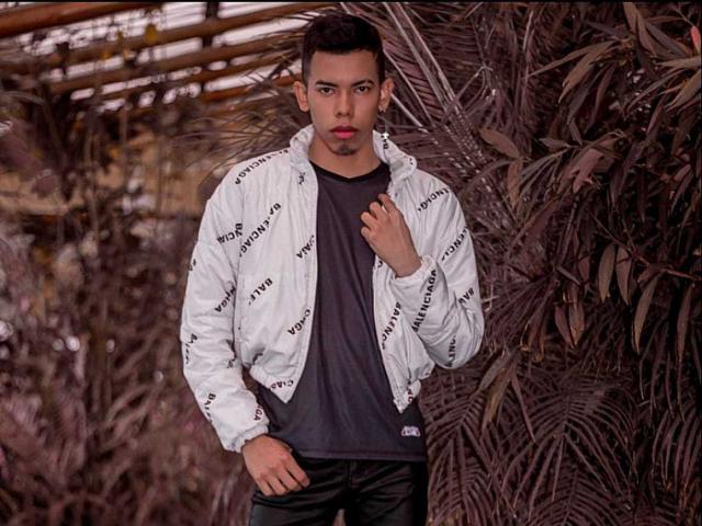 LianStonne with short dark hair, serious expression, wearing a white "VANS" jacket, dark shirt, and black pants, standing against leafy plants, hand on jacket.
