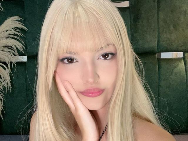 Blonde IrisWithmore with straight hair, brown eyes, and pink lipstick, touching her cheek with her hand, showing off her pretty face and soft smile.