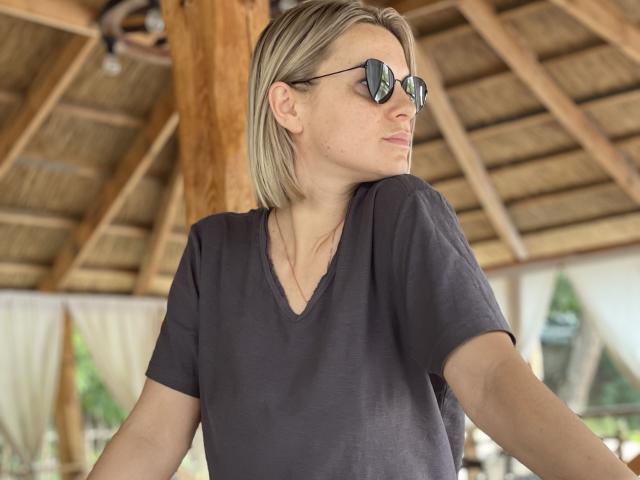 BelleLisaG has blonde hair, wears black sunglasses, and a loose grey V-neck shirt, standing with a relaxed, side-profile gaze in a wooden, thatched-roof structure.