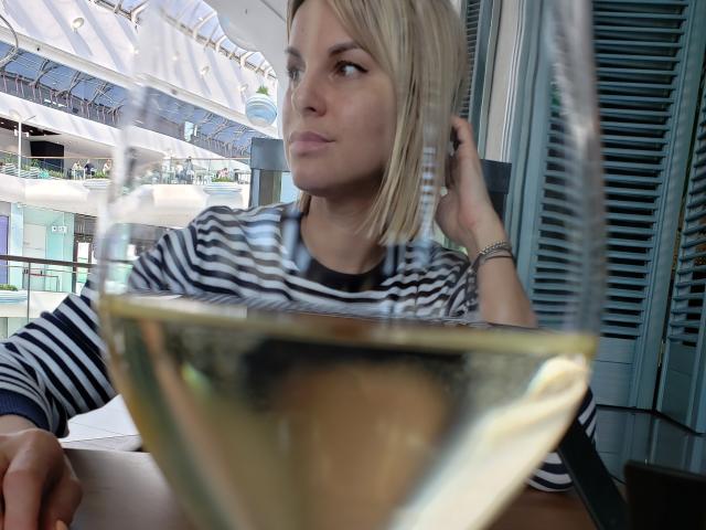 BelleLisaG with blonde bob, wearing striped shirt, looking bored, hand in hair, glass of white wine in foreground, blue shutters, indoor setting, slightly blurred, serious expression.
