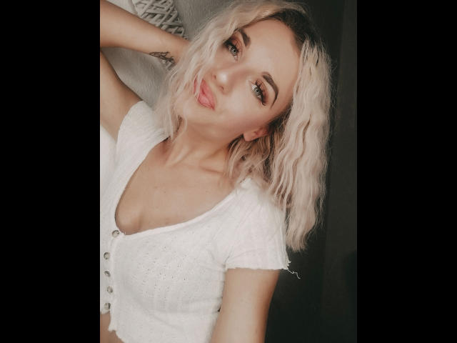 JayJayPink-hot with blonde wavy hair, pouty lips, and white button-up top showing tits, arms raised, smiling, and looking at camera. Soft light, light skin, subtle makeup.