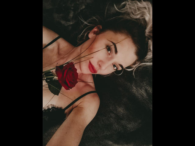 JayJayPink-hot with red rose in mouth, lying on dark fur, black lingerie, hoop earrings, red lipstick, blonde curls, and thin black wires, looking up with seductive eyes.