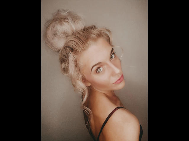 JayJayPink-hot with blonde hair in a messy bun, green eyes, and a slight smile, wearing a black strap, looking over her shoulder. Her tits are barely visible.