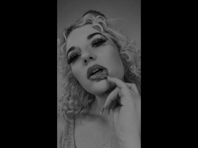 JayJayPink-hot has curly blonde hair, full eyebrows, and is licking her finger with her tongue out. She's looking up with half-closed eyes, wearing dark lipstick. Black-and-white photo.