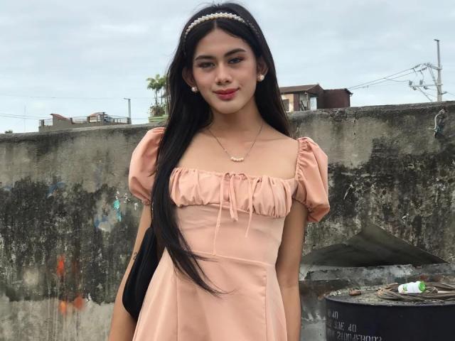 YurieFresh with long black hair, wearing a pink dress, pearl headband, and necklace, standing in front of a dirty wall with a trash can, smiling. Her tits are small.
