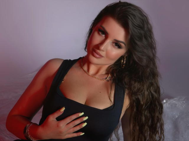 JessieAmore with long brown hair, wearing a black tank top, gold necklace, and yellow nails, covering her large, rounded tits with one hand. She has a cute, sexy smile.