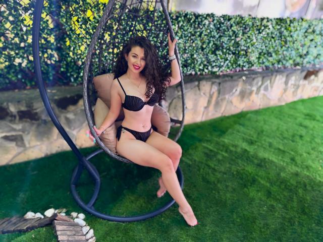 AnaAnais with curly black hair, wearing black lingerie, sits in a hanging chair, showing her tits and ass, on green grass, smiling with red lipstick.