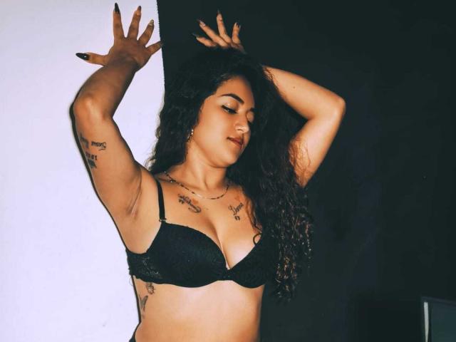 NicoleMartins with long curly hair, black bra, tattoos on arms and chest, arms raised, eyes closed, dark nail polish, light skin, confident pose, sexy, attractive.
