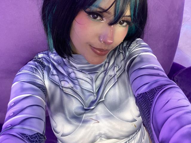 CarlaMiceli with blue and black hair, wearing a shiny silver cosplay outfit, has a nose ring, and is smiling at the camera, showing her tits.