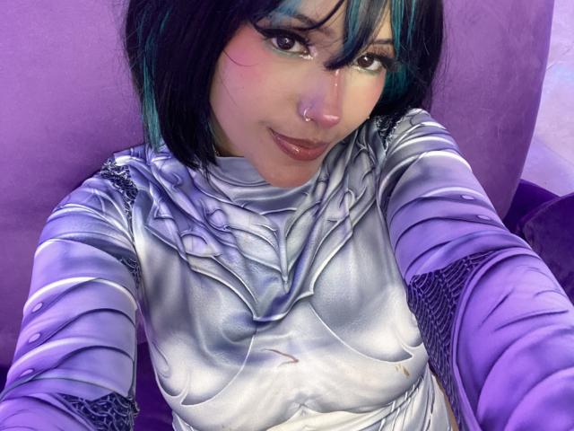 CarlaMiceli with dark blue-green hair, wearing a shiny purple and silver armor-like top, has a nose ring, and is smiling at the camera, showing her tits.