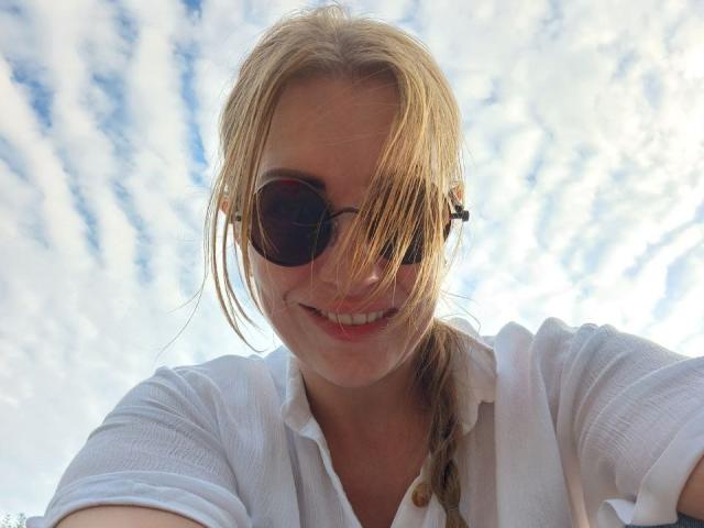 Blonde BlackMango with sunglasses, white shirt, smiling, hair in braid, sky with clouds in background, taking selfie, subtle wind in hair, light skin, cute, relaxed vibe.