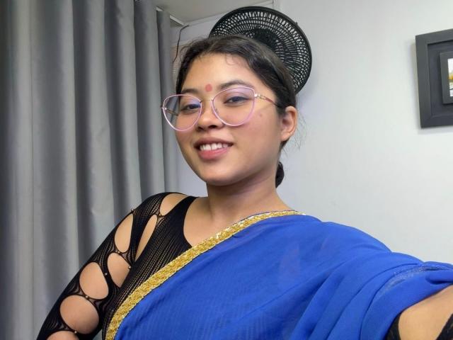 ShailaYojana with glasses, blue sari, black netted top, gold trim, and black headpiece, smiling at camera, showing small red bindi on forehead, brown eyes, cute.