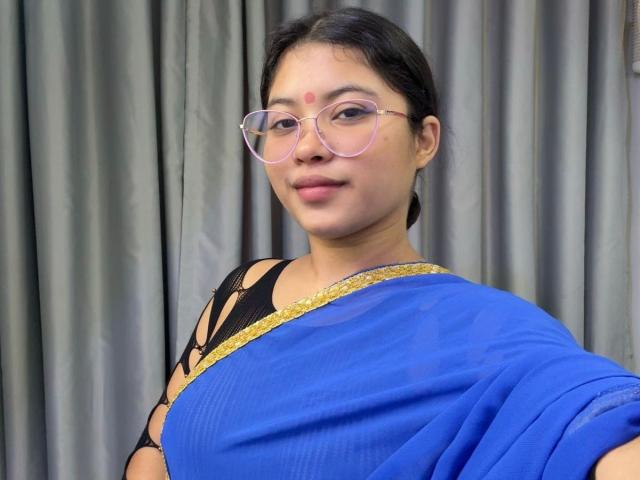 ShailaYojana with pink glasses, blue sari, black bra, and gold trim, smiling, has a red dot on her forehead, dark hair, and a cute face, holding the camera.