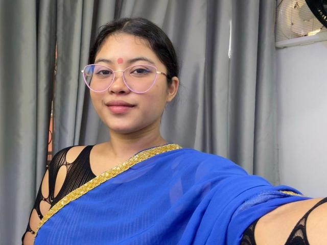 ShailaYojana with glasses, blue sari, black lace, gold trim, and a red bindi, smiling, showing her tits through the sari's cutouts. She has medium brown skin.