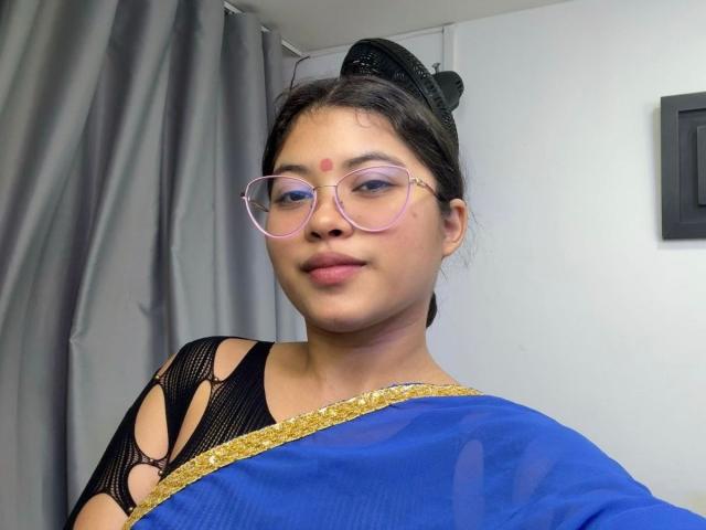 ShailaYojana with pink glasses, brown hair in black bun, blue sari with gold trim, black mesh top, red dot on forehead, smiling, soft light, gray curtain background.