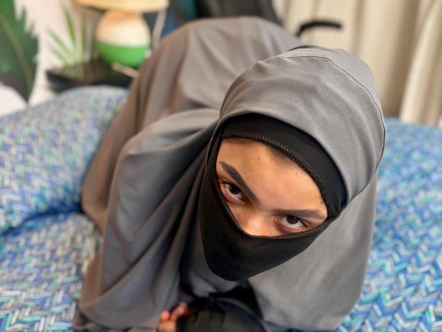 HotMalatha on all fours, wearing a grey hijab and black mask, exposing her brown eyes. Her ass is up, and she's on a blue-patterned bed.
