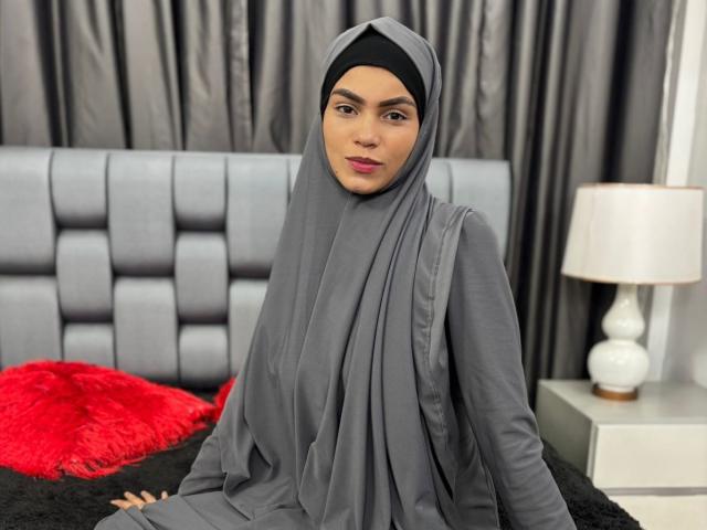 HotMalatha with gray hijab and dress, sitting on black bed, red fuzzy pillow, gray headboard, and white lamp, looking at camera, pink lipstick, dark eyebrows, sexy.