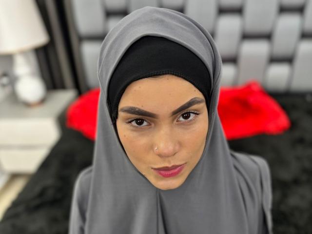 HotMalatha with a gray hijab, black undercap, brown eyes, pink lips, nose piercing, and a serious expression, sitting on a black bed with a red pillow.