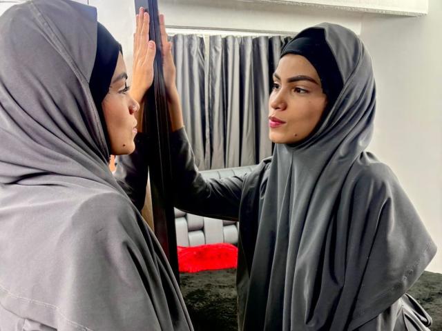 HotMalatha in a gray hijab, standing in front of a mirror, touching it with her right hand, showing off her red lipstick and dark eyebrows, looking sexy.