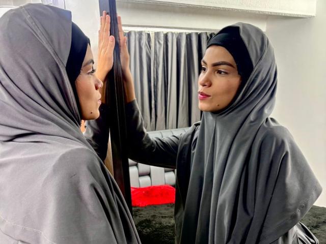 HotMalatha with grey hijab, reflection in mirror, right hand on mirror, looking at herself, pink lipstick, black eyebrows, dark skin, black hair, grey hijab, white wall background.