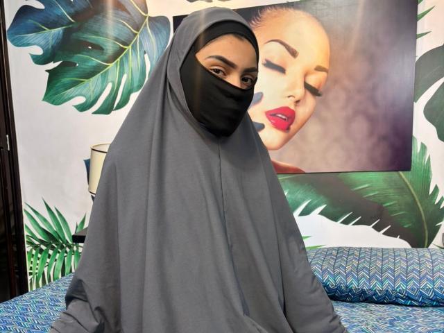HotMalatha with a gray hijab, black mask covering her mouth and nose, brown eyes looking at camera, red lipstick visible, blue patterned pillow in background.