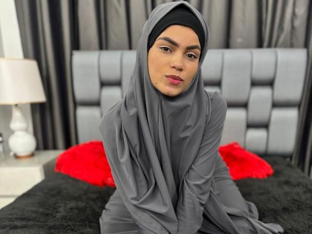HotMalatha with gray hijab, sitting on black bed, gray headscarf covering her tits, black eyebrows, pink lipstick, red fluffy pillow, white lamp, gray tufted headboard in background.