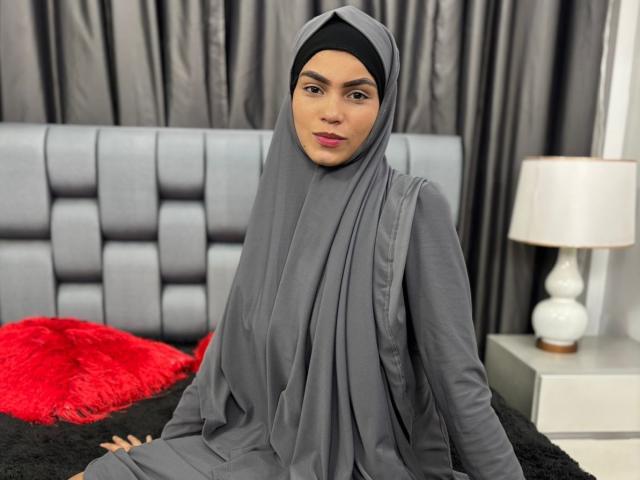 HotMalatha with gray hijab and long dress, sitting on black bed, gray headboard, red fluffy pillow, white nightstand with lamp. Her ass is slightly visible under dress.